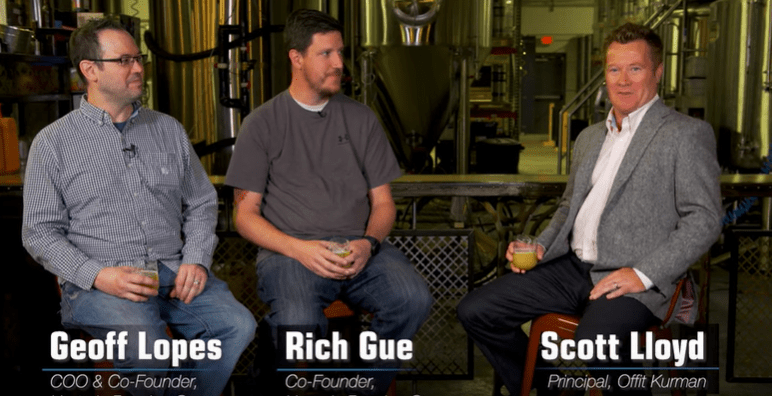 Video pic of Geoff Lopes and Rich Gue, Co-Founders of Hysteria Brewing Company interviewed by Scott Lloyd. 3 Men talking with each other and holding a glass filled with beer in their hands sitting in a distilling factory