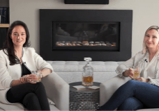 Image of Rachel Mech & Emily Shank sitting in front of an electric fireplace with drinks in their hands
