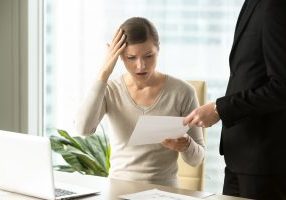 Puzzled businesswoman sitting at desk being handed a lawsuit document by a man in a black suit