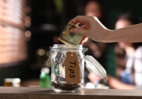 Woman putting 20 dollar bill into a glass tip jar on wooden table indoors, closeup