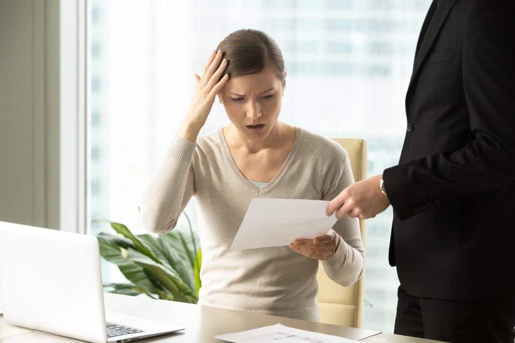 Puzzled businesswoman sitting at desk being handed a lawsuit document by a man in a black suit