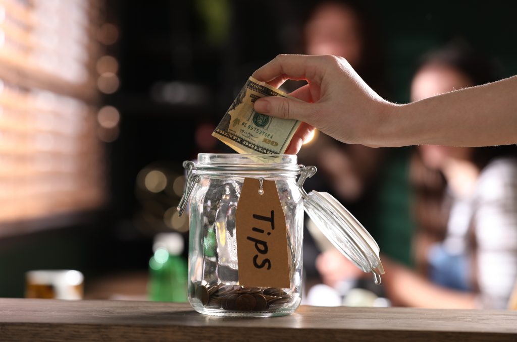Woman putting 20 dollar bill into a glass tip jar on wooden table indoors, closeup