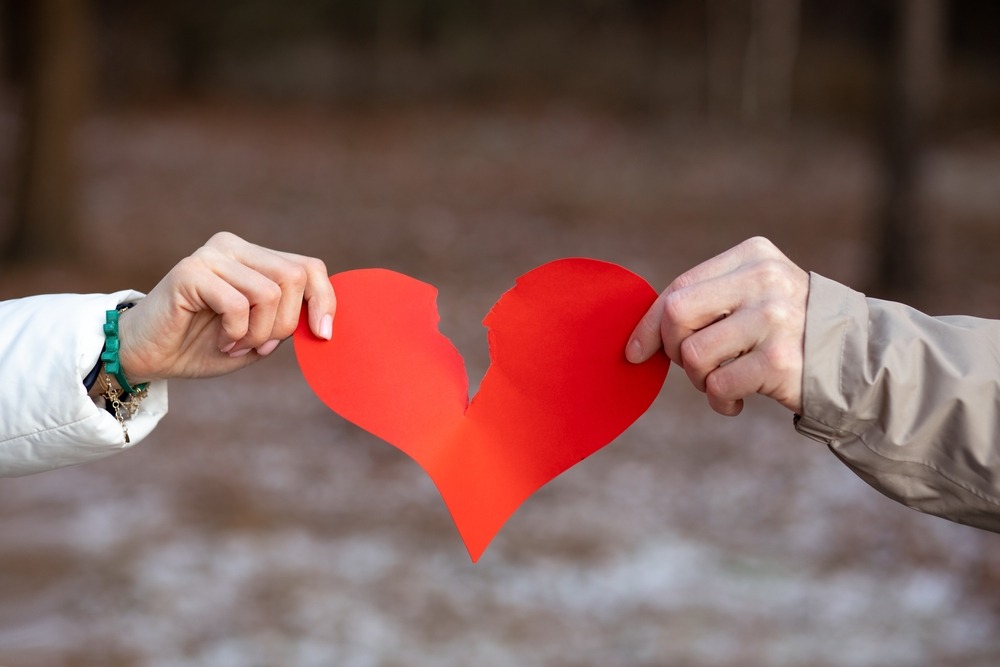 Two hands, male and female, tear a red paper heart against background of winter forest.