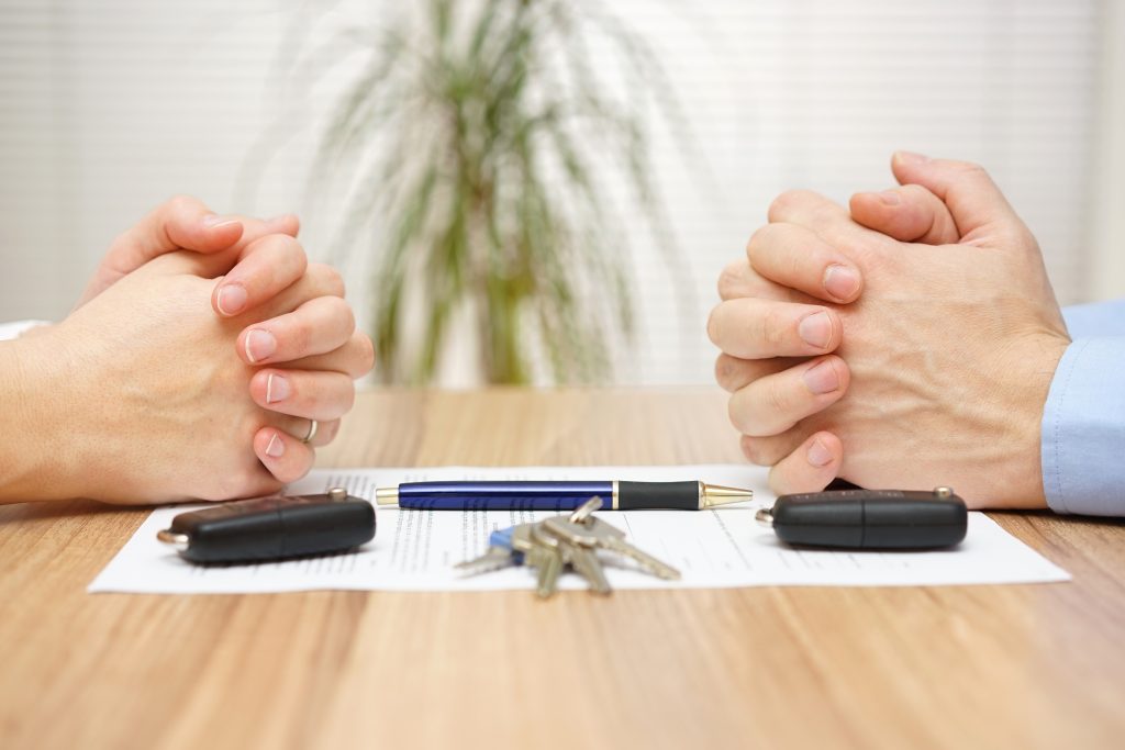 close up of a man's hands and a woman's hands sitting across a desk from each other with a paper, pen and car keys in between them.